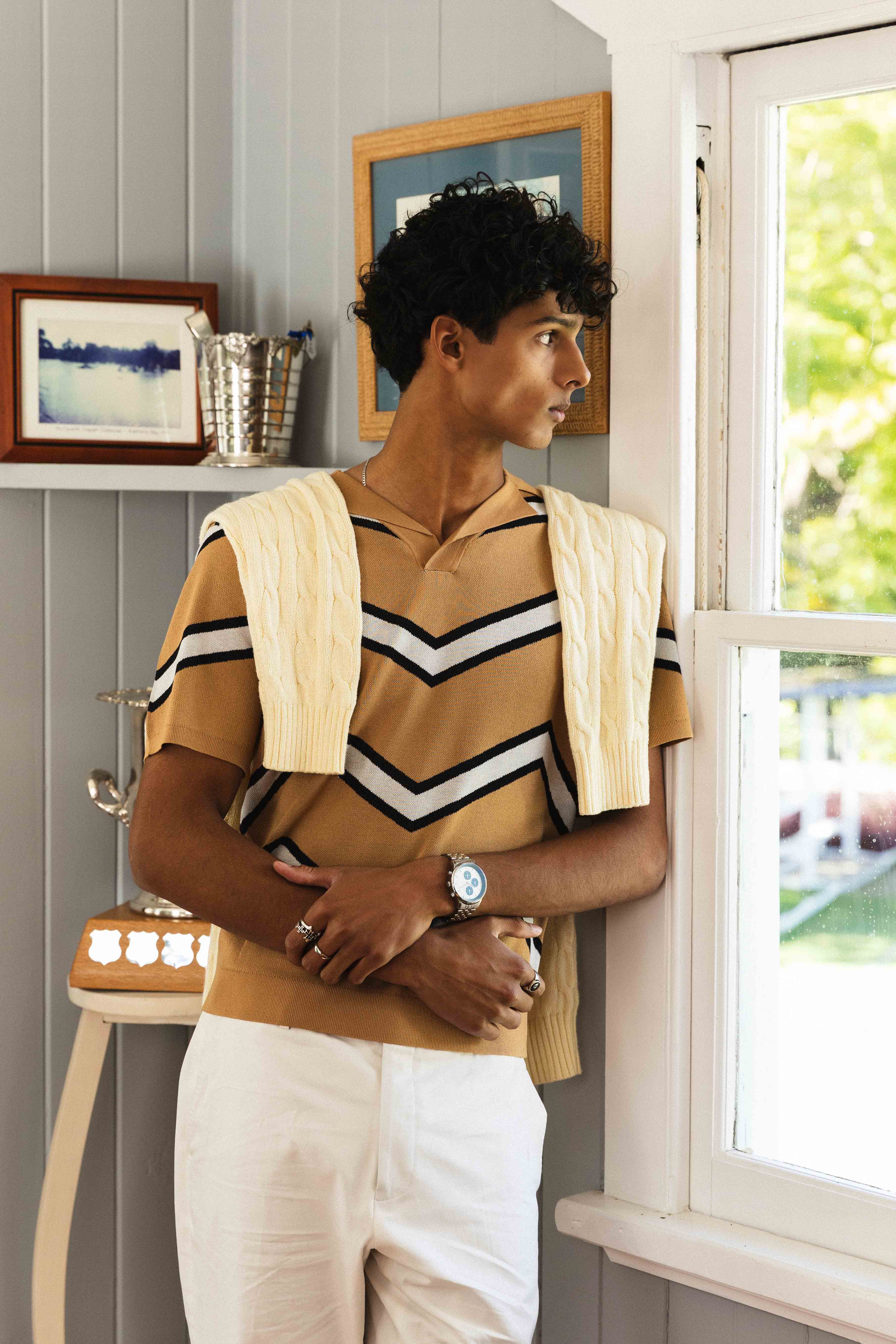 Woman standing by a window wearing a striped shirt and white pants, with a neutral background.