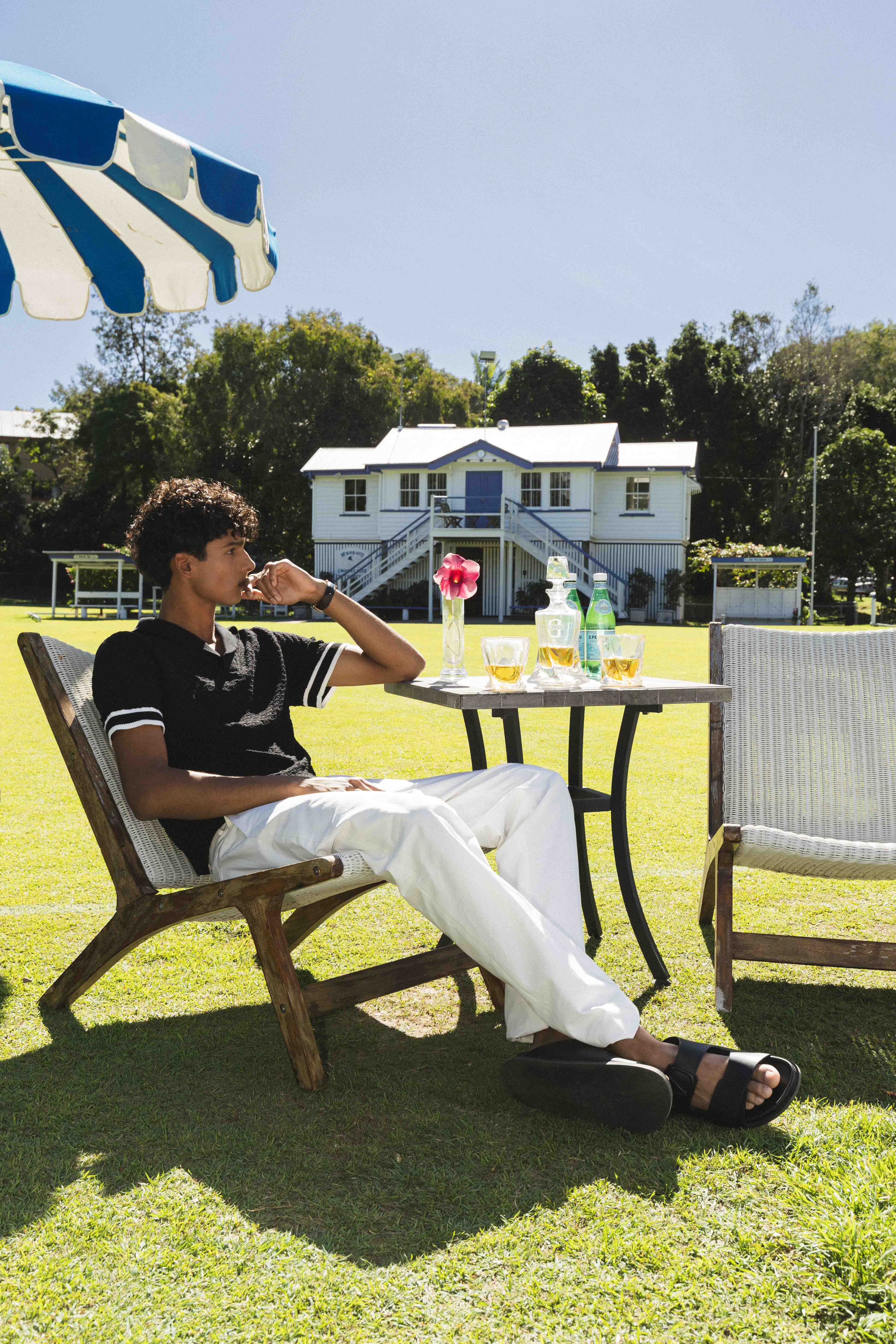 Man sitting outdoors on a lawn with a table and chairs, enjoying a drink.