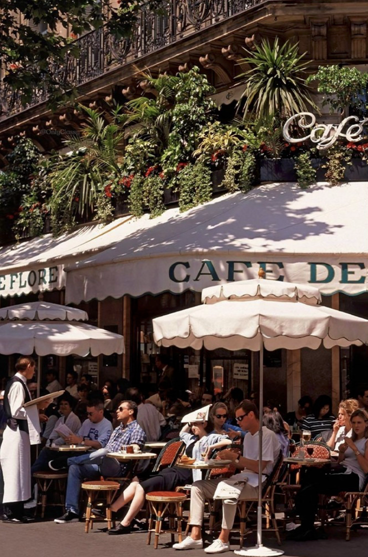 Outdoor cafe scene with people sitting under white umbrellas, surrounded by greenery and a building facade.