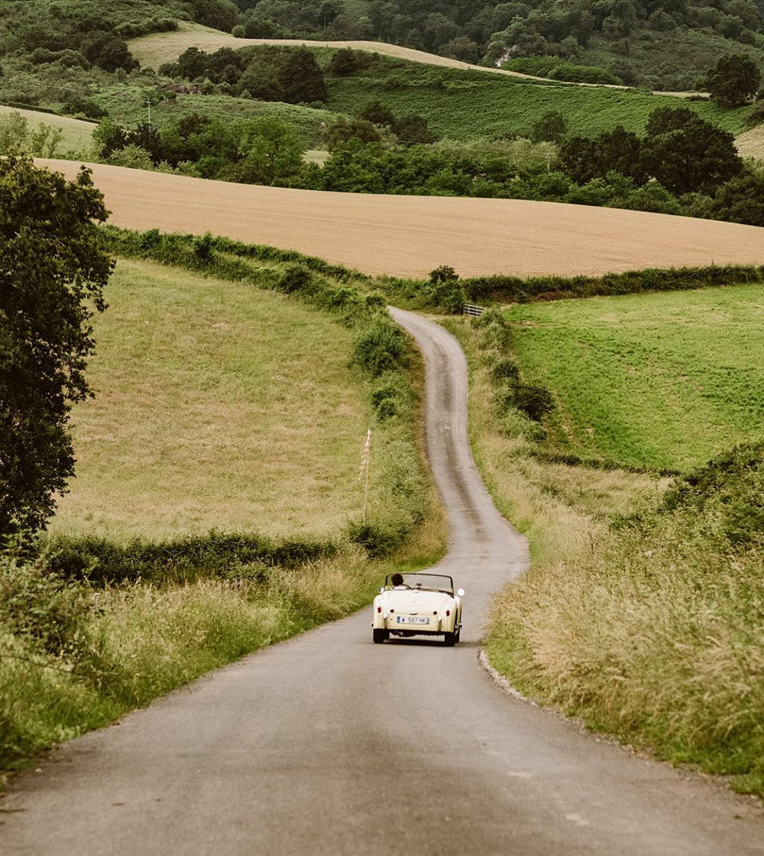 Car driving down a winding country road with green fields on either side