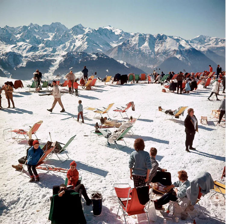 People enjoying a sunny day on a snowy mountain with colorful chairs and mountains in the background.